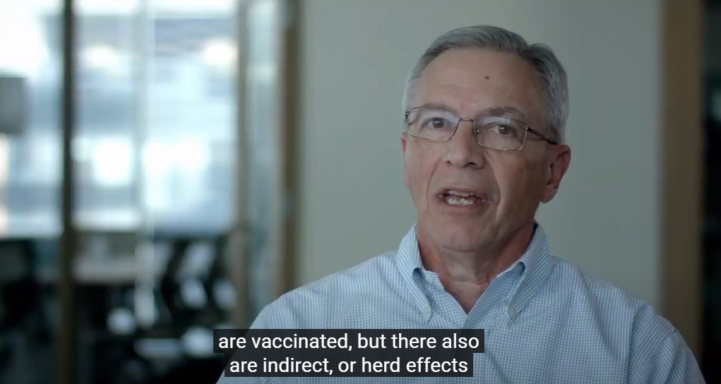 A man with glasses and short gray hair is speaking indoors. He is wearing a light blue, checkered dress shirt. The background is blurred, showing a window and some office furniture. Subtitles on the screen read: "are vaccinated, but there also are indirect, or herd effects".