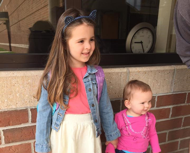 Two young girls are standing in front of a brick wall and window. The older girl is wearing a denim jacket, cat ear headband, and a backpack. The younger girl is in a pink top. A round clock is visible in the background through the window.