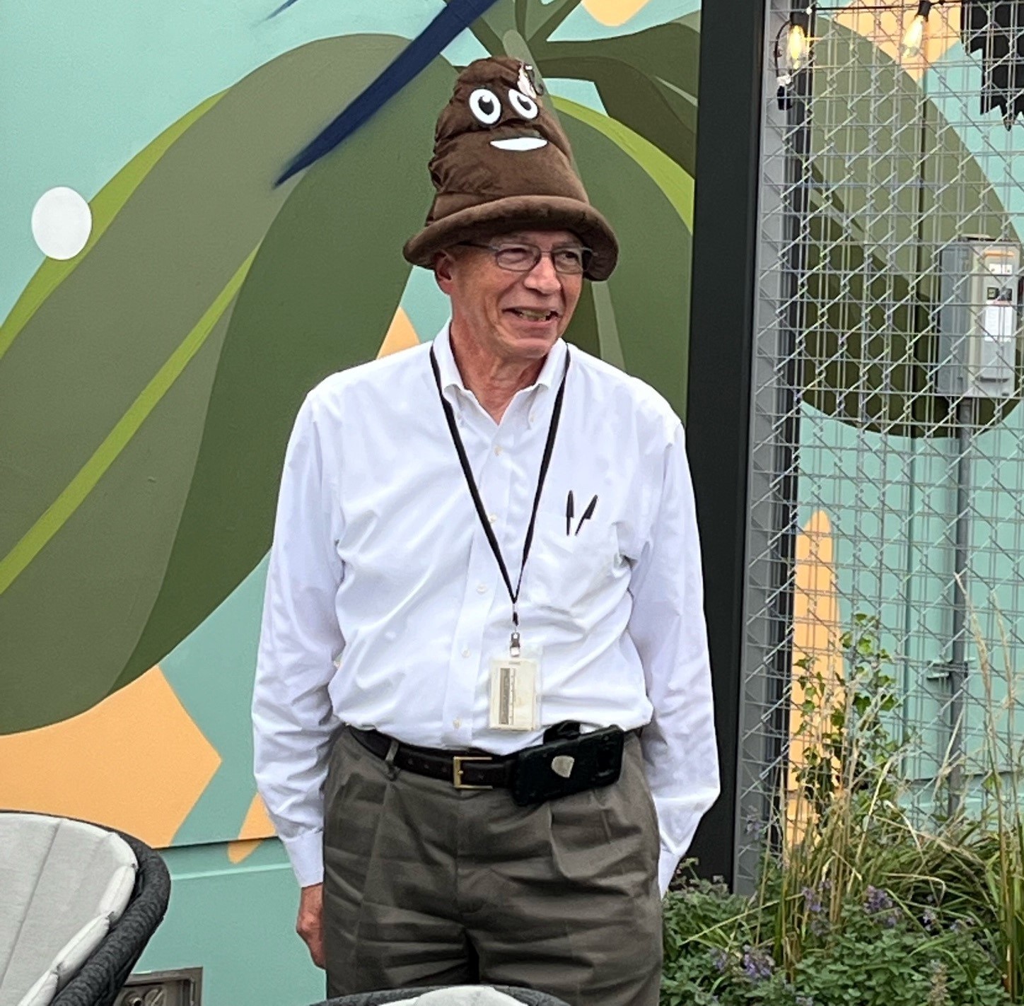 An elderly man wearing glasses and a white shirt stands outdoors. He is smiling and has a brown poop emoji hat on his head. The background features a fence and a colorful mural with green and blue tones. There are plants and a chair nearby.