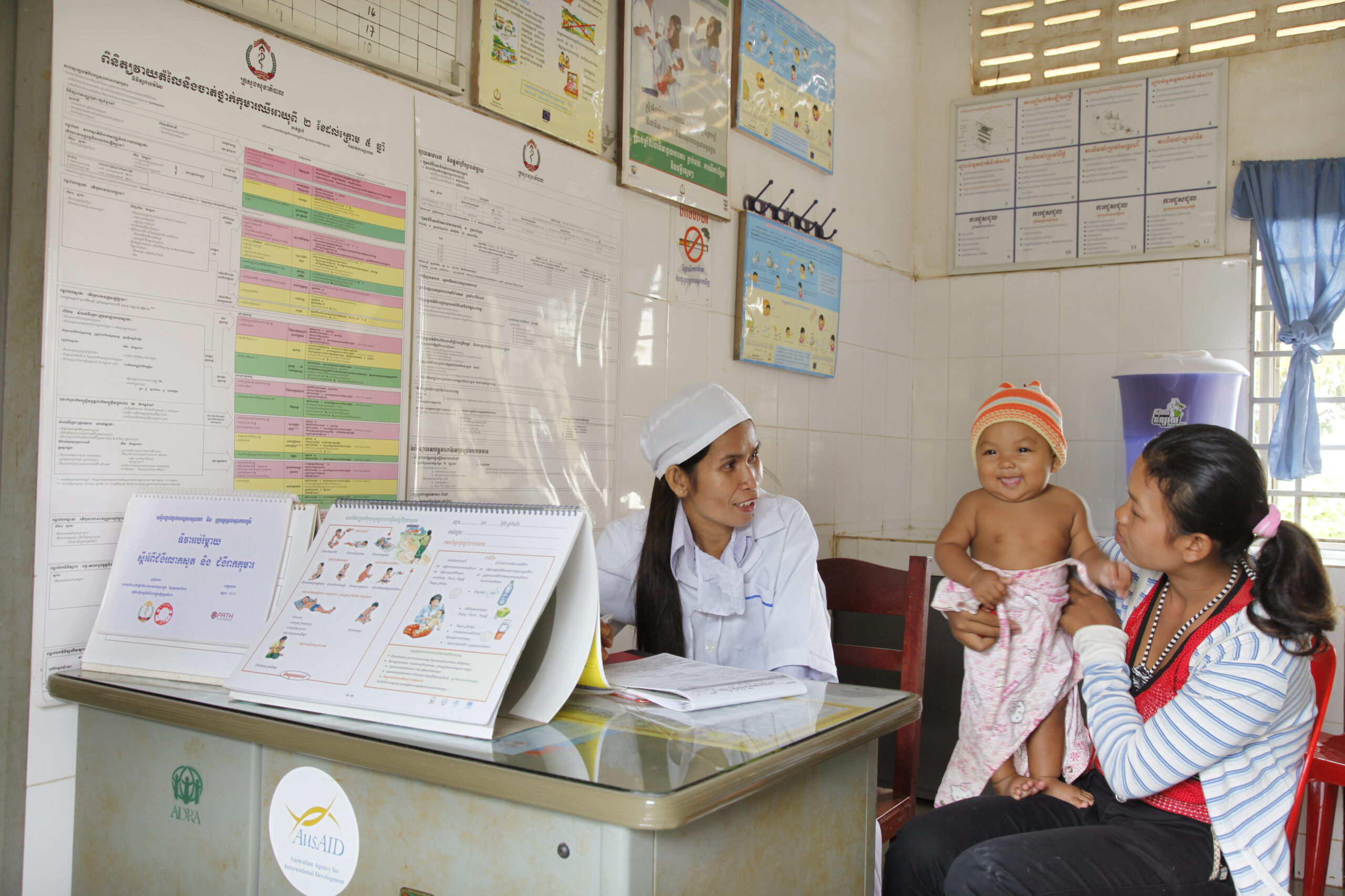 A health worker sits at a desk, speaking with a mother holding her baby in a clinic. Charts and informational posters are displayed on the wall. The mother is seated, smiling at her baby, who is wrapped in a blanket and wearing a striped hat.