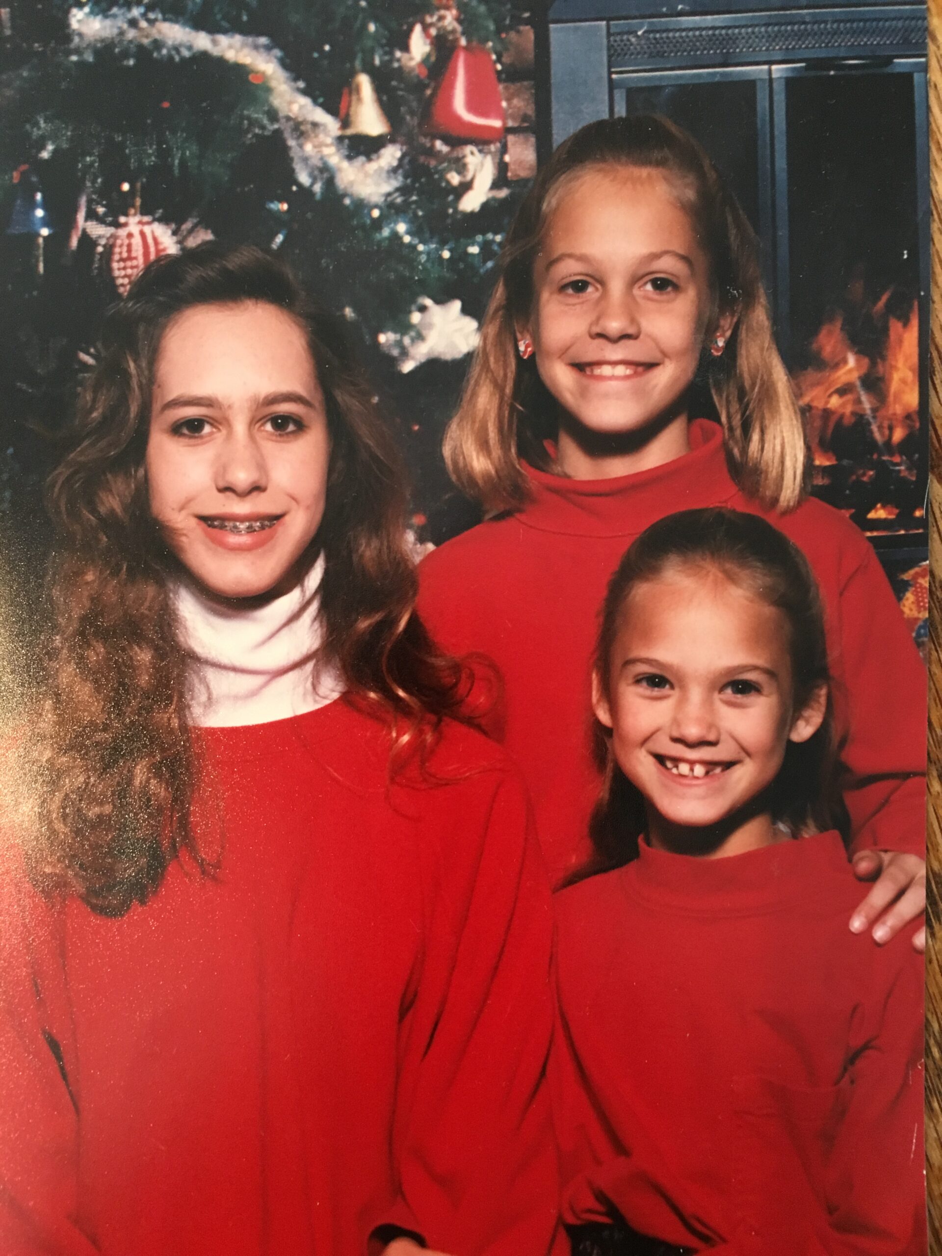 Three young girls in red sweaters pose in front of a decorated Christmas tree and a fireplace. The girl on the left has light brown hair, the one in the middle has blonde hair in a half-up style, and the smallest girl on the right has a big smile and blonde hair.