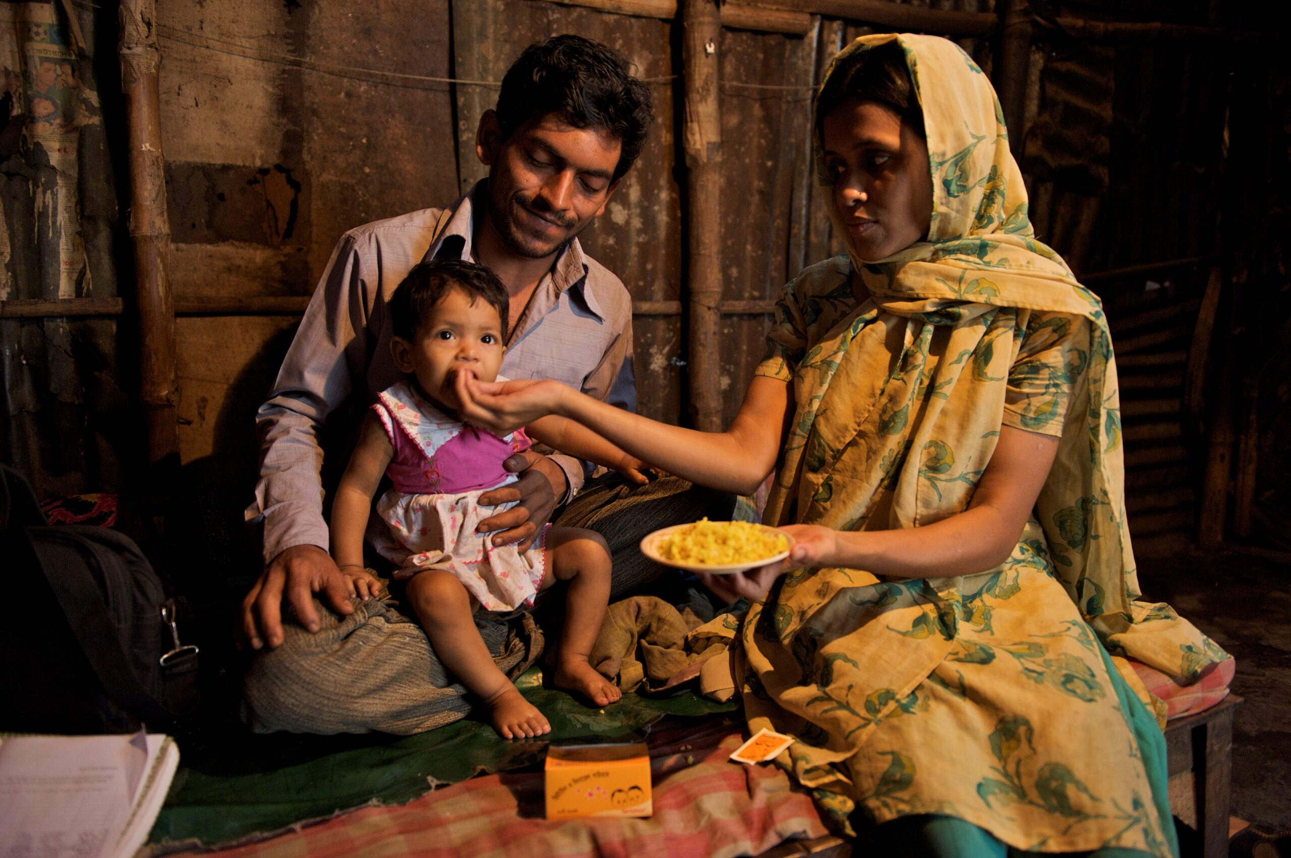 A man and woman sit closely together, the man holding a child on his lap. The woman, dressed in a yellow patterned sari, feeds the child from a bowl of food. They sit on the floor in a rustic setting, with bamboo and wooden walls. Some personal items are around them.