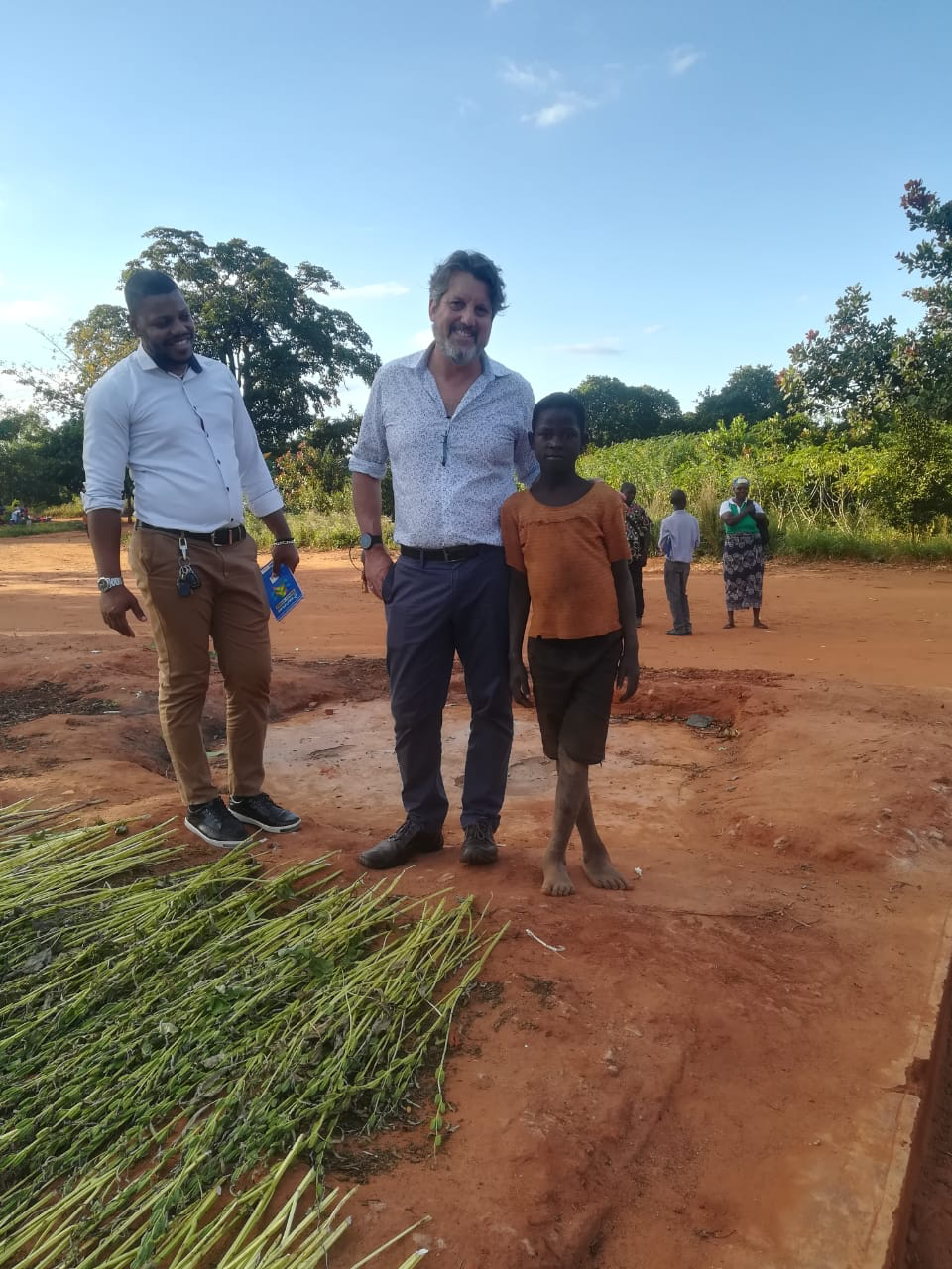 Three people are standing outdoors on bare, reddish soil near some green plants laid out on the ground. An adult in a white shirt and khaki pants stands to the left. An adult in a gray shirt and dark pants stands in the center with their arm around a child in an orange shirt and black shorts. Other people are visible in the background among greenery.