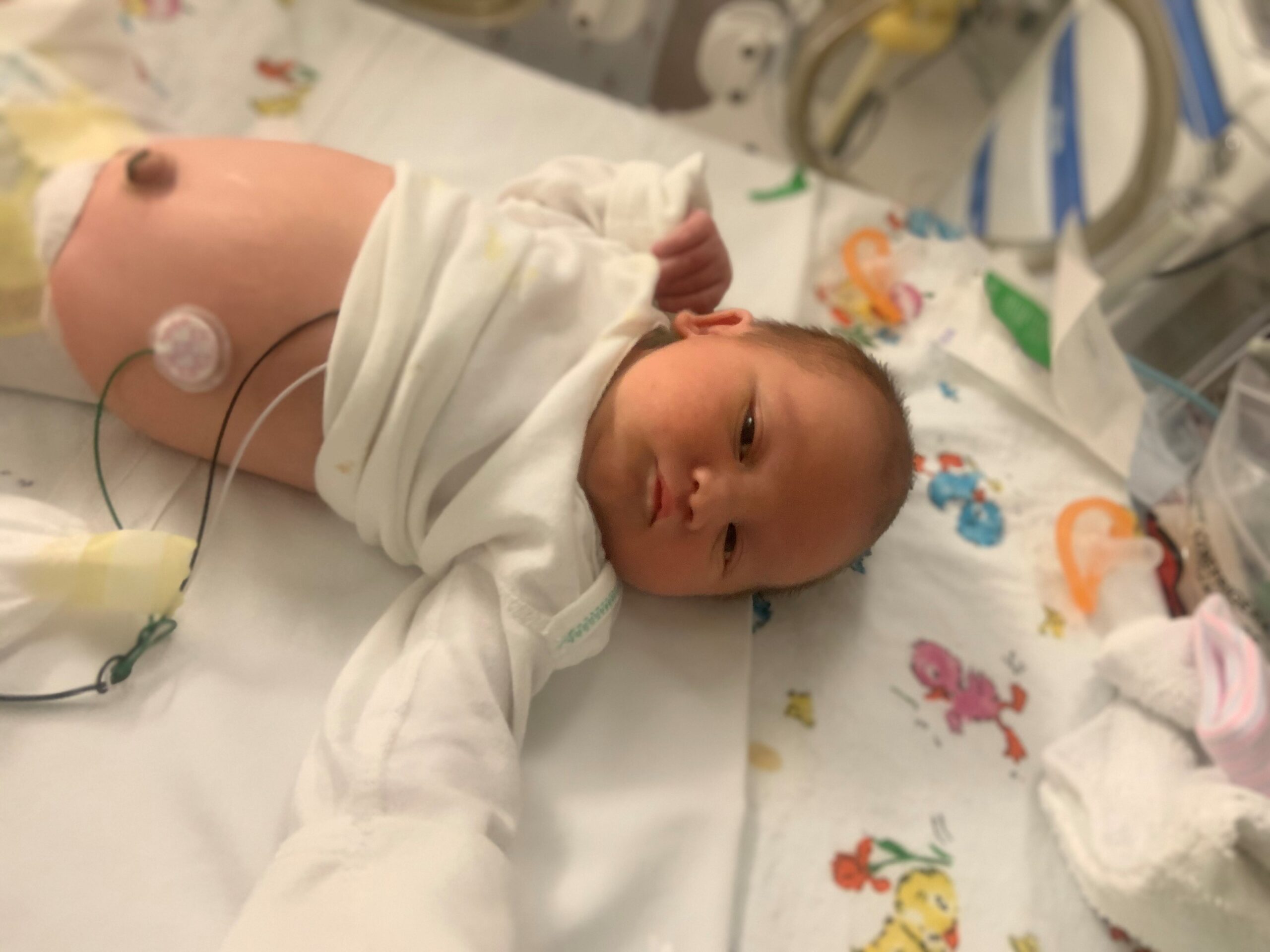 A newborn baby lies in a hospital crib, wearing a white onesie. Medical tubes are attached to the baby. The background shows colorful printed blankets and medical equipment.