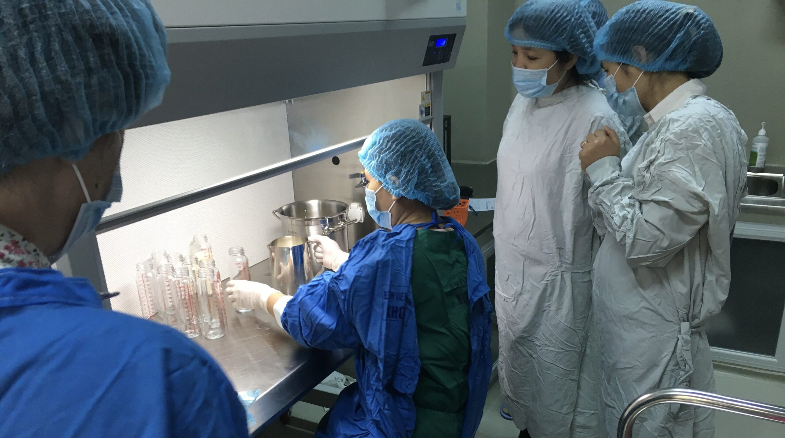 A group of people in a laboratory setting, wearing protective gear including blue hairnets, masks, white lab coats, and gloves. One person is seated and working with lab equipment while the others stand close by, observing the process.