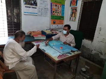 A man wearing a mask sits at a desk, while an elderly man sitting across from him appears to be providing some documents. The office has posters on the wall and various files and objects on the desk. The room has barred windows.