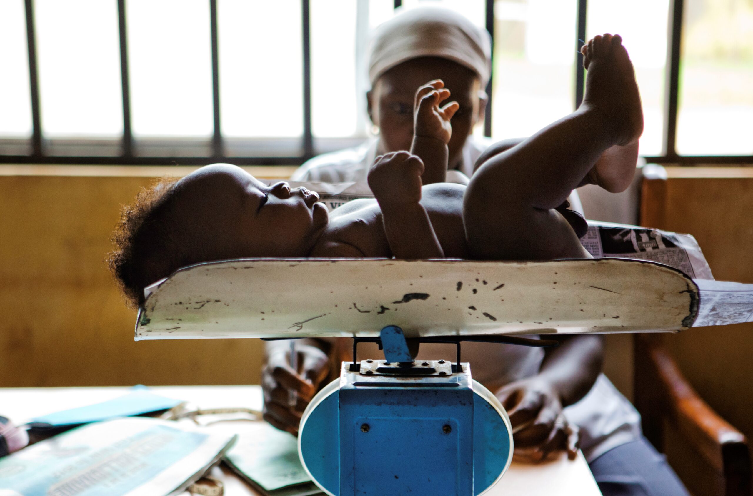 A baby lies on a weighing scale with arms and legs raised, while an adult sits behind the scale, reading a document. The background shows window bars and a dimly lit room. The scene appears to be in a clinic or healthcare setting.