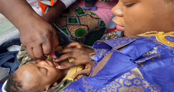 A caregiver administers oral medication to a baby held by a woman dressed in a blue and gold patterned outfit. The baby lies calmly with eyes closed, surrounded by colorful fabrics.