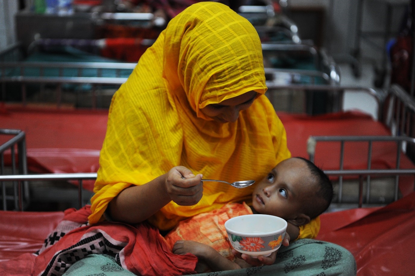 A woman in a yellow headscarf is seated, holding a spoon and feeding a small child in her lap. They are in a room with several beds. The child looks up at her while she holds a bowl in one hand. The atmosphere is warm and caring.