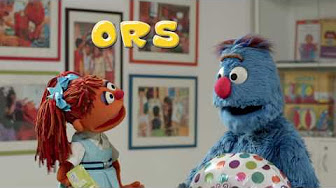 Two colorful muppet characters converse in a room decorated with framed pictures. One character, with red hair, wears a blue dress and holds a booklet. The other, a blue furry character with a red nose, holds a large, colorful egg. The text "ORS" appears above them.