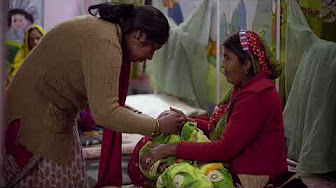 A woman leans over and tends to a baby wrapped in a colorful blanket, cradled by another woman sitting on a bed. The setting appears to be a modest room with curtains and other beds, suggesting a hospital or care facility.