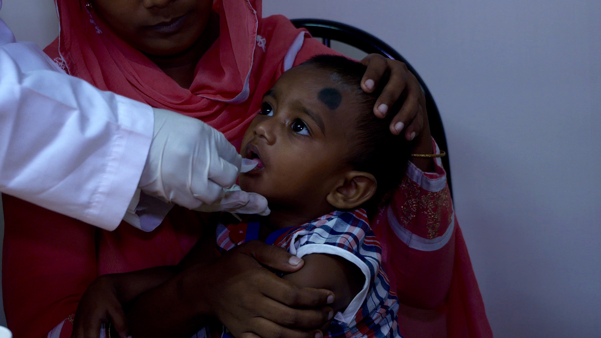 A young child receives an oral vaccine administered by a healthcare professional wearing gloves. The child is sitting in an adult's lap, and the adult holds the child's head steady. Both appear to be inside a clinic.