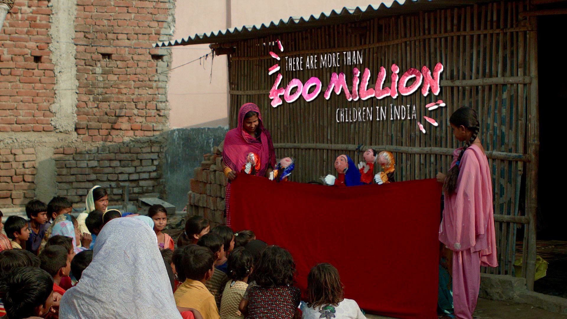 A group of children sit outdoors watching a puppet show performed by three women using a red cloth. Text above reads, "There are more than 400 million children in India!" The scene is set against a rustic building with a mix of brick and bamboo walls.