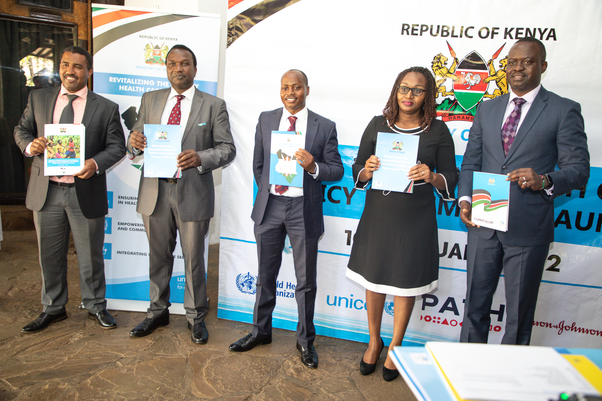 A group of five individuals, dressed in business attire, stand in a row holding documents in front of a banner. The banner bears the Republic of Kenya emblem and mentions a health policy and strategy launch event held in January 2022.