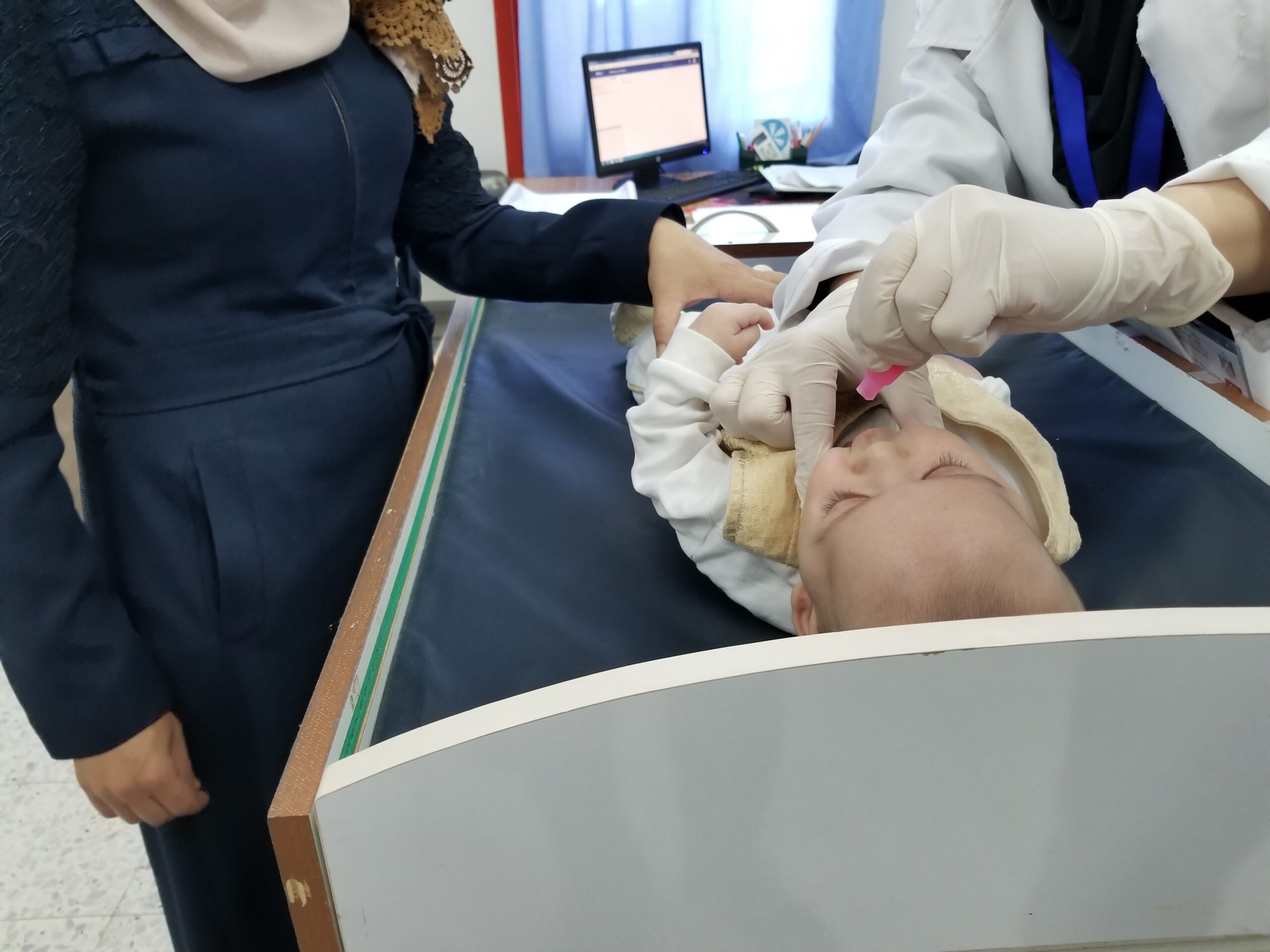 A healthcare worker in a white coat and gloves administers an oral vaccine to a baby lying on an examination table. A woman, possibly the baby's mother, stands beside the table, gently holding the baby's arm. A computer screen is visible in the background.