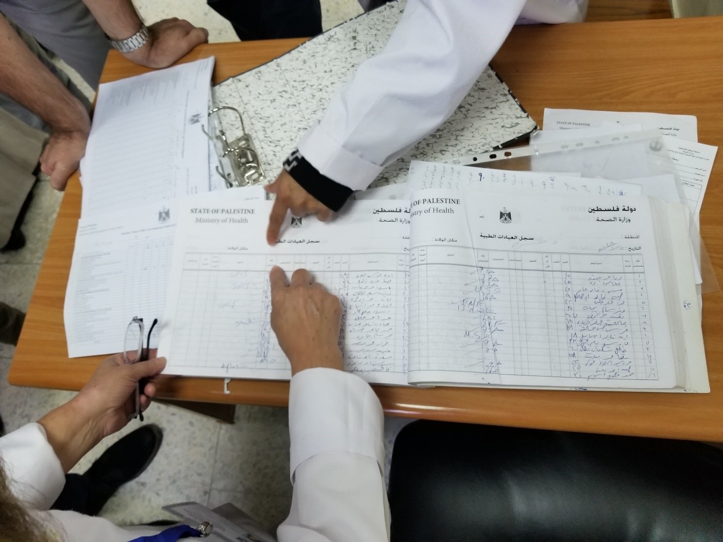 Hands of several individuals examining official documents labeled "Ministry of Health" on a wooden table, with papers and a clipboard nearby. The documents appear to contain lists written in Arabic script.