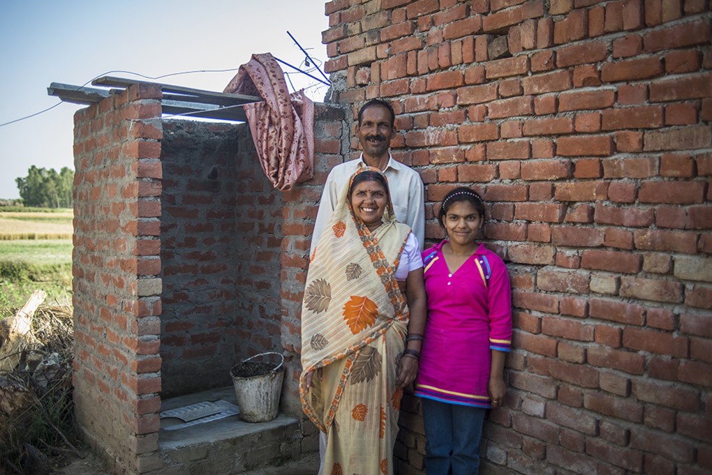 A family of three stands smiling in front of a brick structure on a sunny day. The woman wears a patterned saree, the man is in a light shirt, and the young girl is in a bright pink outfit. A bucket is on the ground nearby.