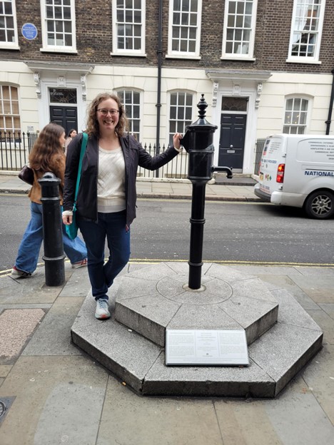 A woman with curly hair, glasses, and a black jacket is standing next to a black water pump on a hexagonal stone base with a plaque. She is smiling and holds the pump with one hand. The background shows a street, brick buildings, another person, and a parked van.