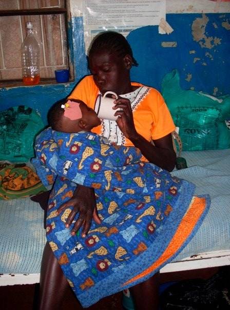 A person wearing an orange shirt is sitting on a bed, holding and feeding a child wrapped in a colorful blanket with animal prints. The child has a bandage on their forehead. The background includes a window, medical supplies, and a blue wall.