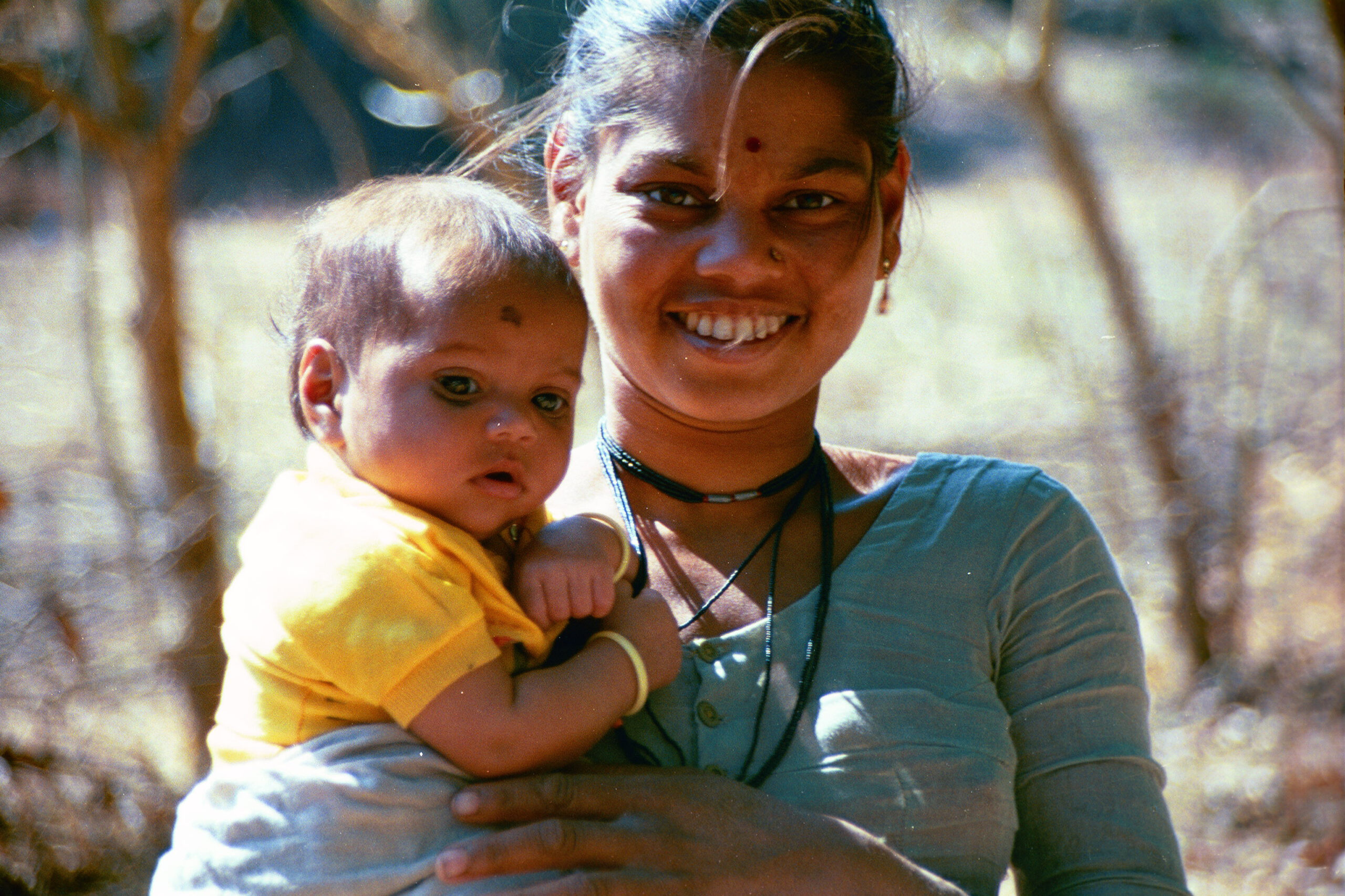 A woman smiles at the camera while holding a baby. The baby is wearing a yellow shirt and has a small mark on its forehead. The woman wears a light blue top, a necklace, and earrings. They are standing outdoors, with trees and blurred natural background.