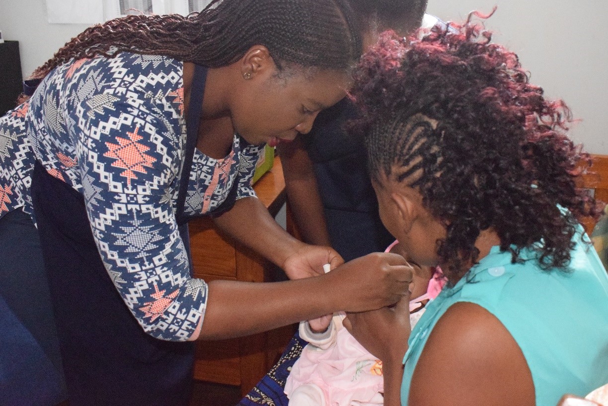 A person wearing a patterned blouse assists another person in a turquoise top with a small task involving a baby dressed in pink, who is cradled between them. The setting appears to be indoors.