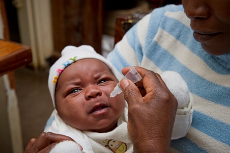 A close-up of a baby receiving an oral vaccine. The baby is wearing a white hat and onesie, while an adult's hand is gently holding the baby's head and administering the vaccine with a dropper. The adult wears a white and blue striped sweater.