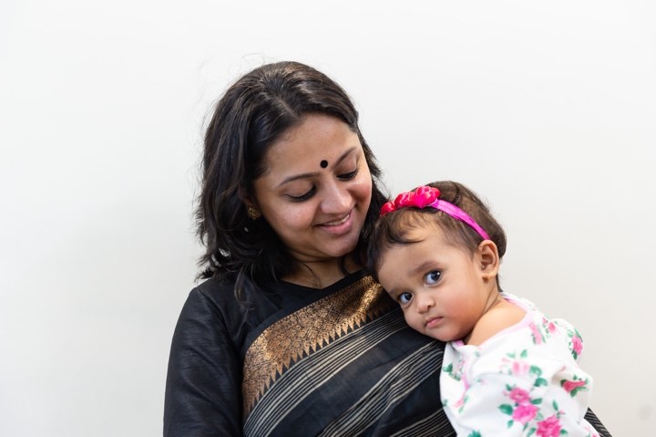 A woman with a black bindi and wearing a black and gold saree is holding a young child dressed in floral clothing. The child, adorned with a pink headband, looks at the camera while the woman gazes affectionately at the child.