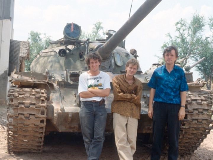 Three individuals are standing in front of a large military tank. The person on the left wears a white T-shirt, the person in the middle is in a brown shirt, and the person on the right is dressed in a blue shirt. The scene appears to be outdoors with trees in the background.