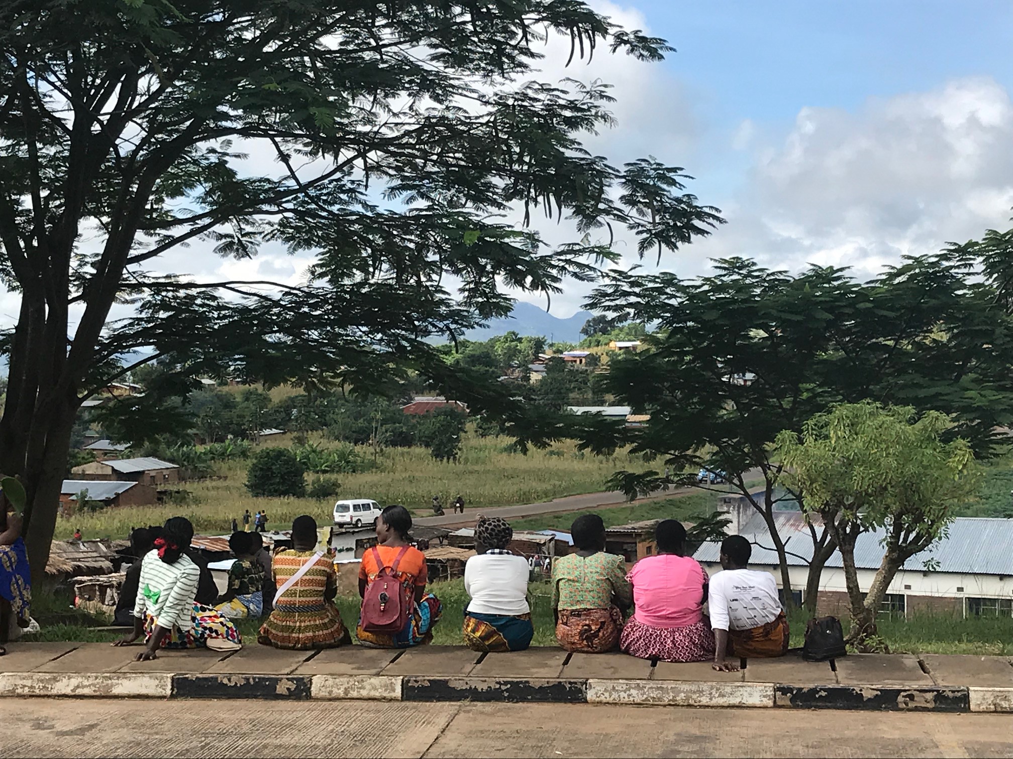 A group of people sit on a concrete path under large trees, looking out over a scenic rural landscape with buildings, a road, and distant mountains under a cloudy sky.