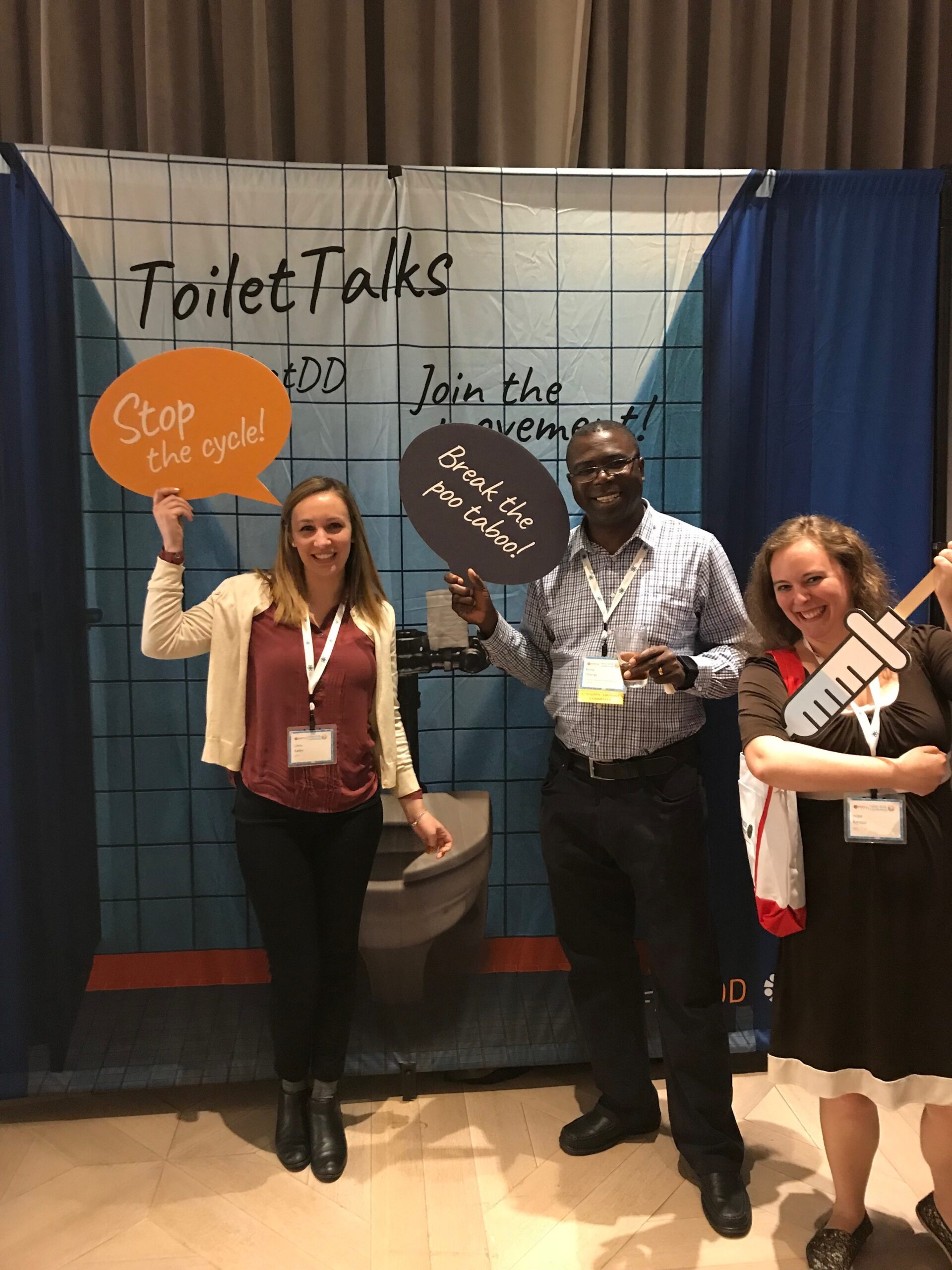 Three people stand in front of a backdrop that reads "Toilet Talks," "Join the movement." They hold speech bubble props with phrases: "Stop the cycle!" "Break the poo taboo!" and a prop shaped like a giant toothbrush. All are smiling and wearing name tags.