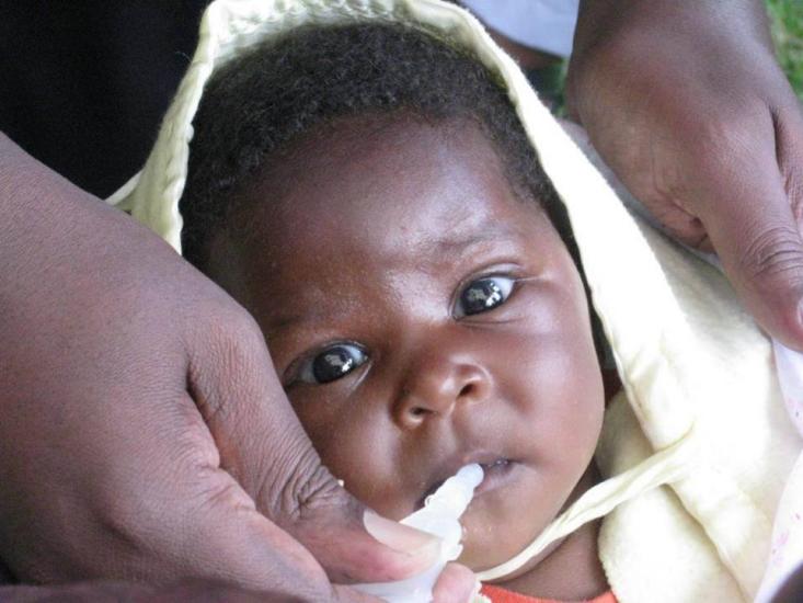 A close-up of an infant with dark skin wearing a light-colored hood and being held by adult hands. The baby is looking up with wide eyes while receiving an oral vaccine. The background is blurred but seems to be outdoors.