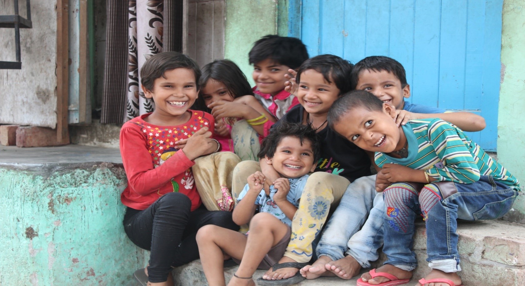 A group of seven children sit and kneel on steps outside a building with a blue door. They are smiling and laughing, wearing colorful clothes. The background includes a brick wall, a curtain, and weathered paint. The scene is joyful and lively.