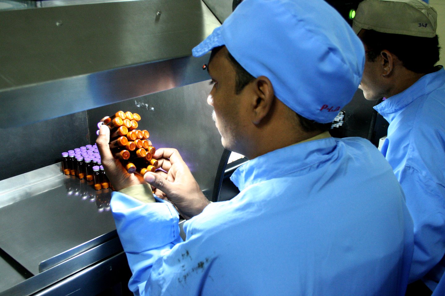 Two workers in blue uniforms inspect a cluster of vaccine vials under a light in a laboratory setting. The focus is on the vials, with attention to detail in the inspection process.