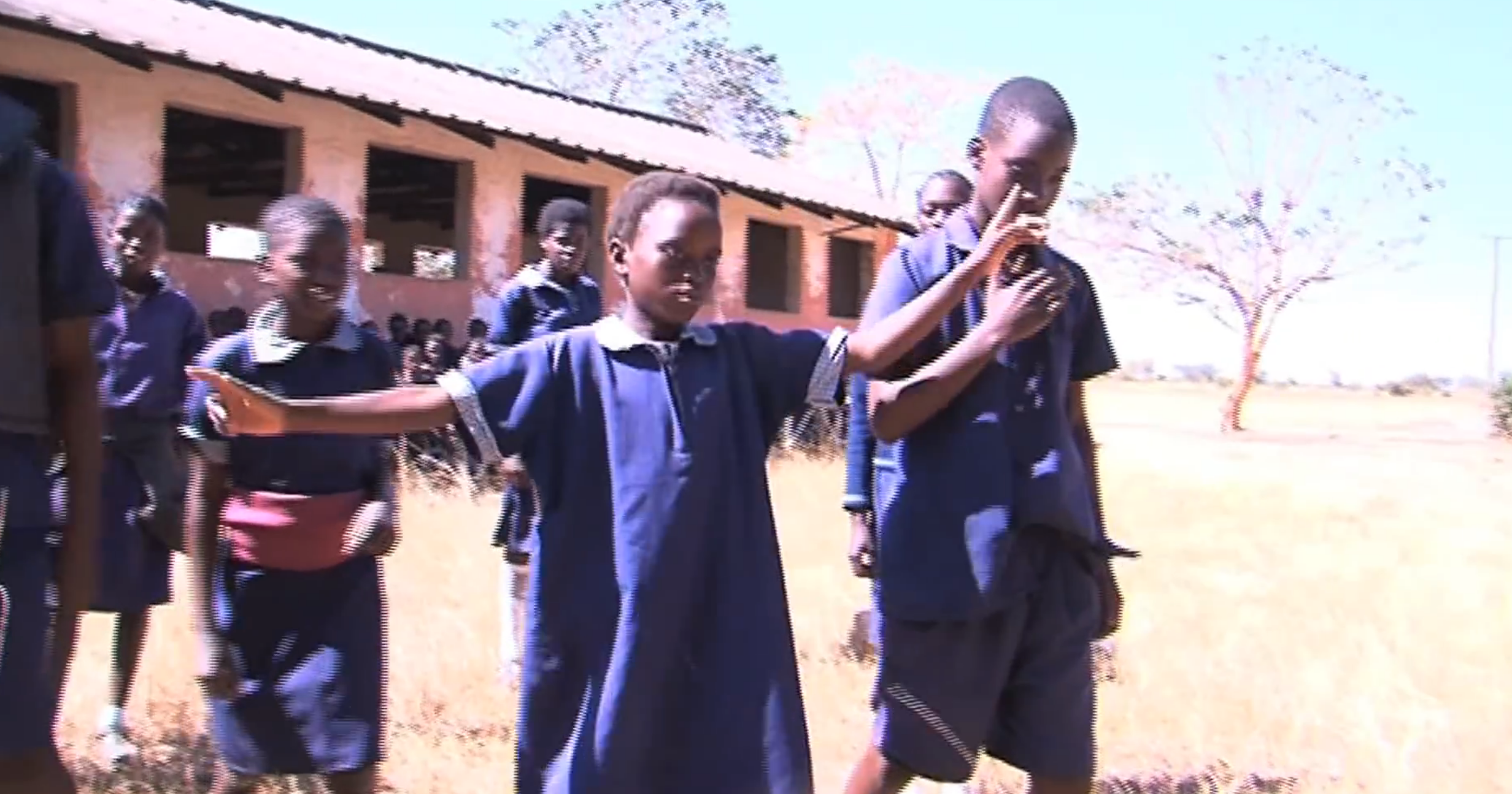 A group of children in blue uniforms walk outside near a school building. One child has arms outstretched as others walk beside them on a grassy area under clear skies.