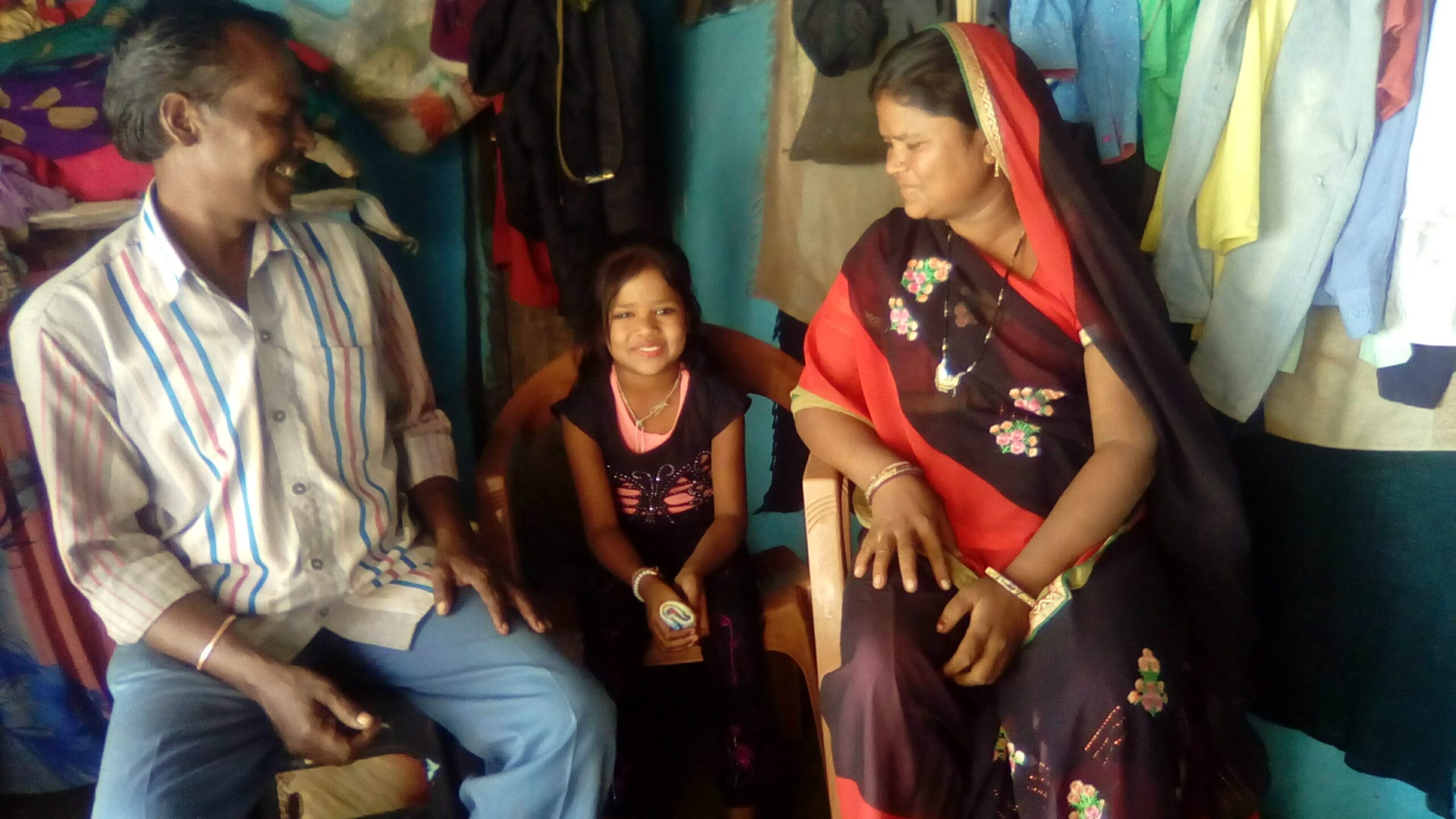 A young girl smiles while sitting between an older man and a woman in a brightly colored room filled with clothes hanging on racks. The man is wearing a striped shirt and blue pants, and the woman is in a black sari with red trim and floral embroidery.