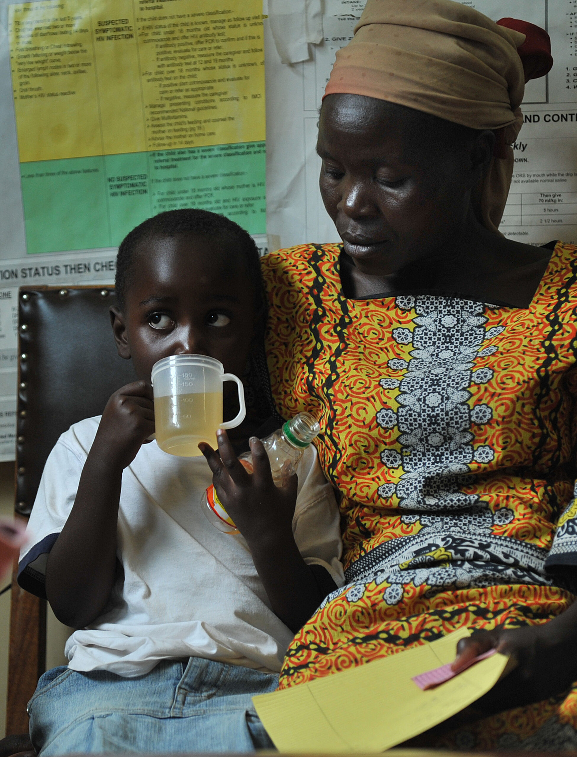 A child drinks from a white cup as a woman in a colorful dress and headscarf holds a green bottle and reads a document. They sit together, surrounded by informational posters on a wall behind them.