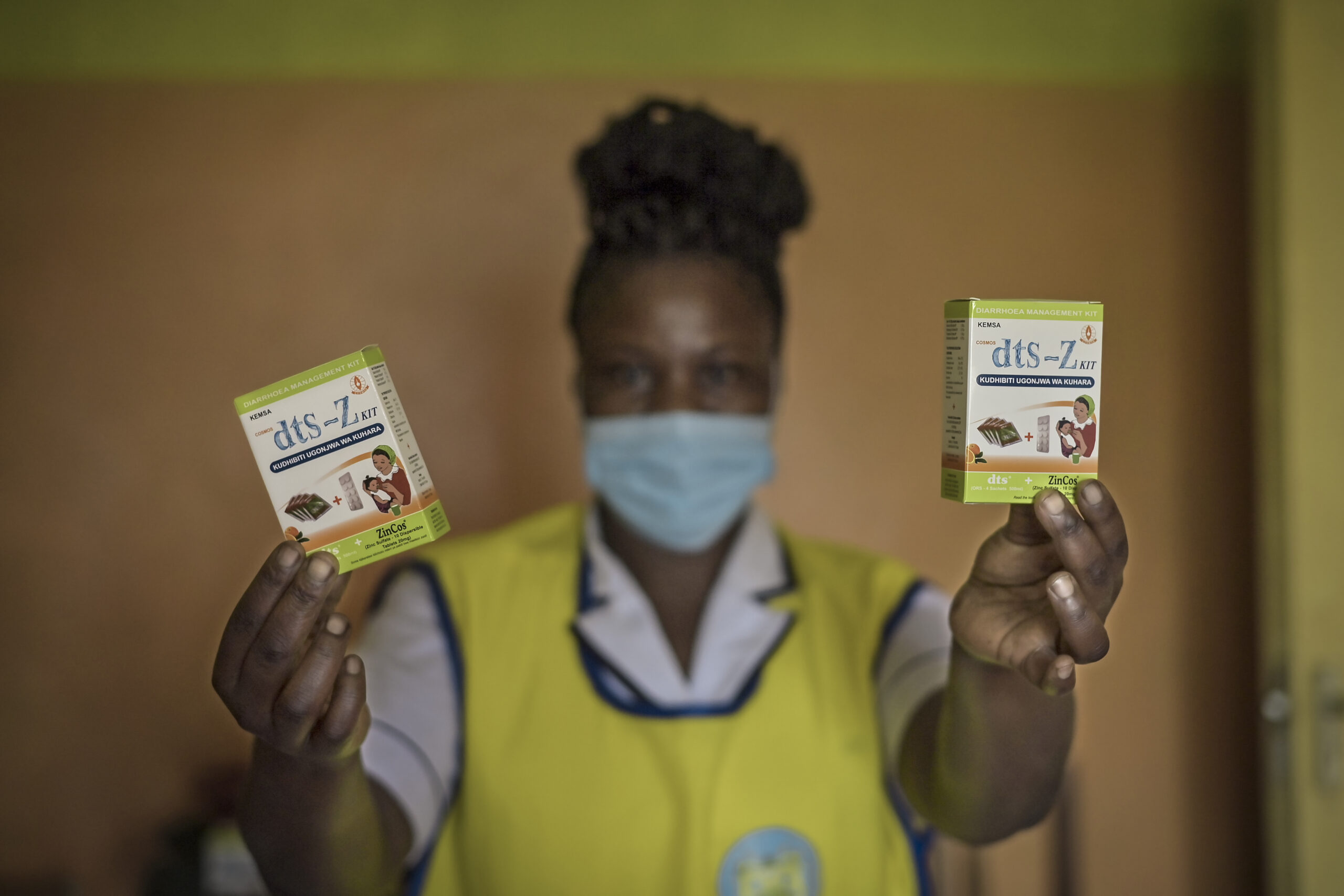 A person wearing a yellow vest, white shirt, and a medical mask holds two small green boxes of medication labeled "dts-Z Luv." Both boxes show images of healthy children. The background is blurred and has a greenish-yellow hue.