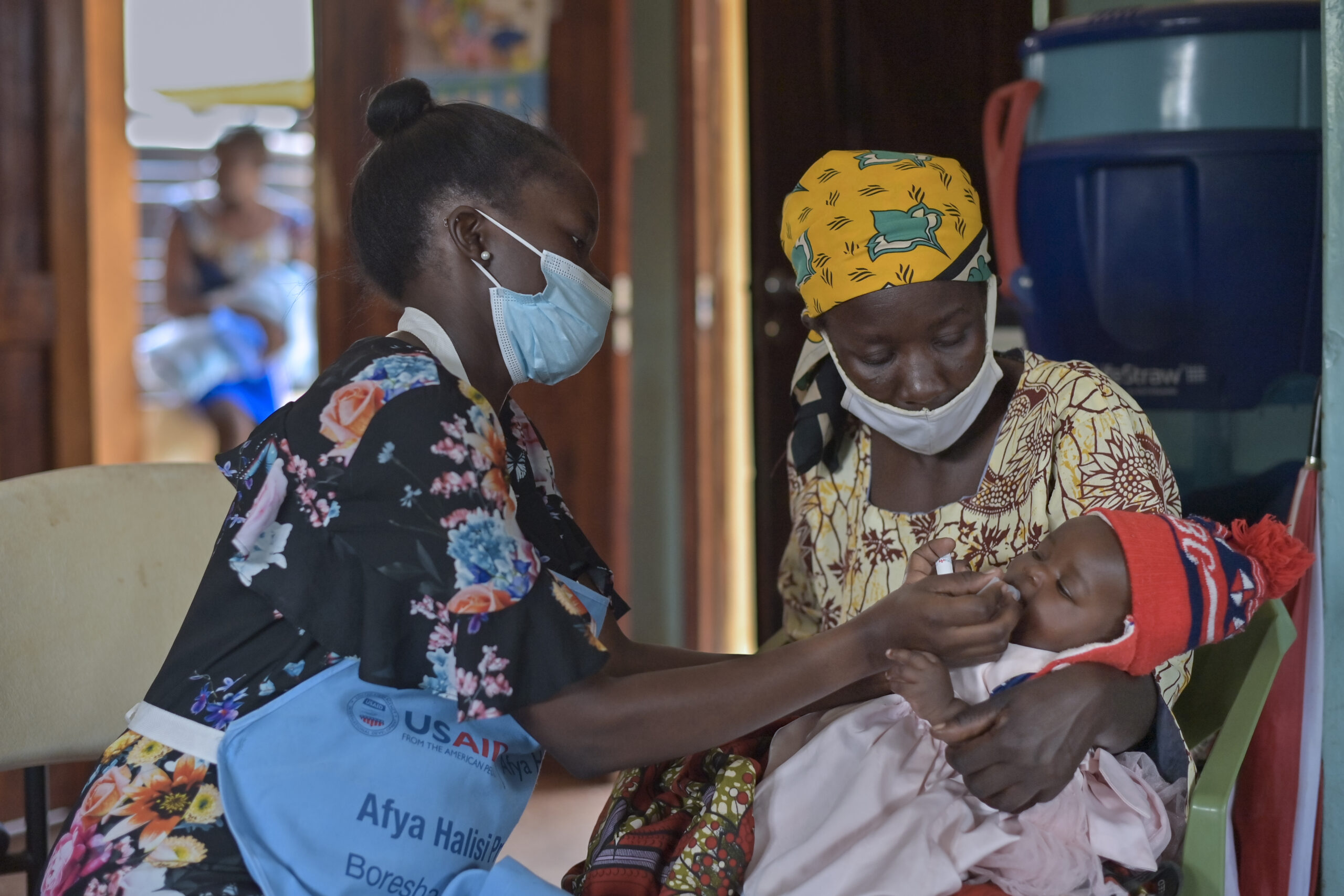 A healthcare worker in a floral print dress administers an oral vaccine to a baby held by a woman wearing a colorful headscarf and patterned clothing. The scene is indoors with medical supplies in the background, emphasizing a healthcare setting.