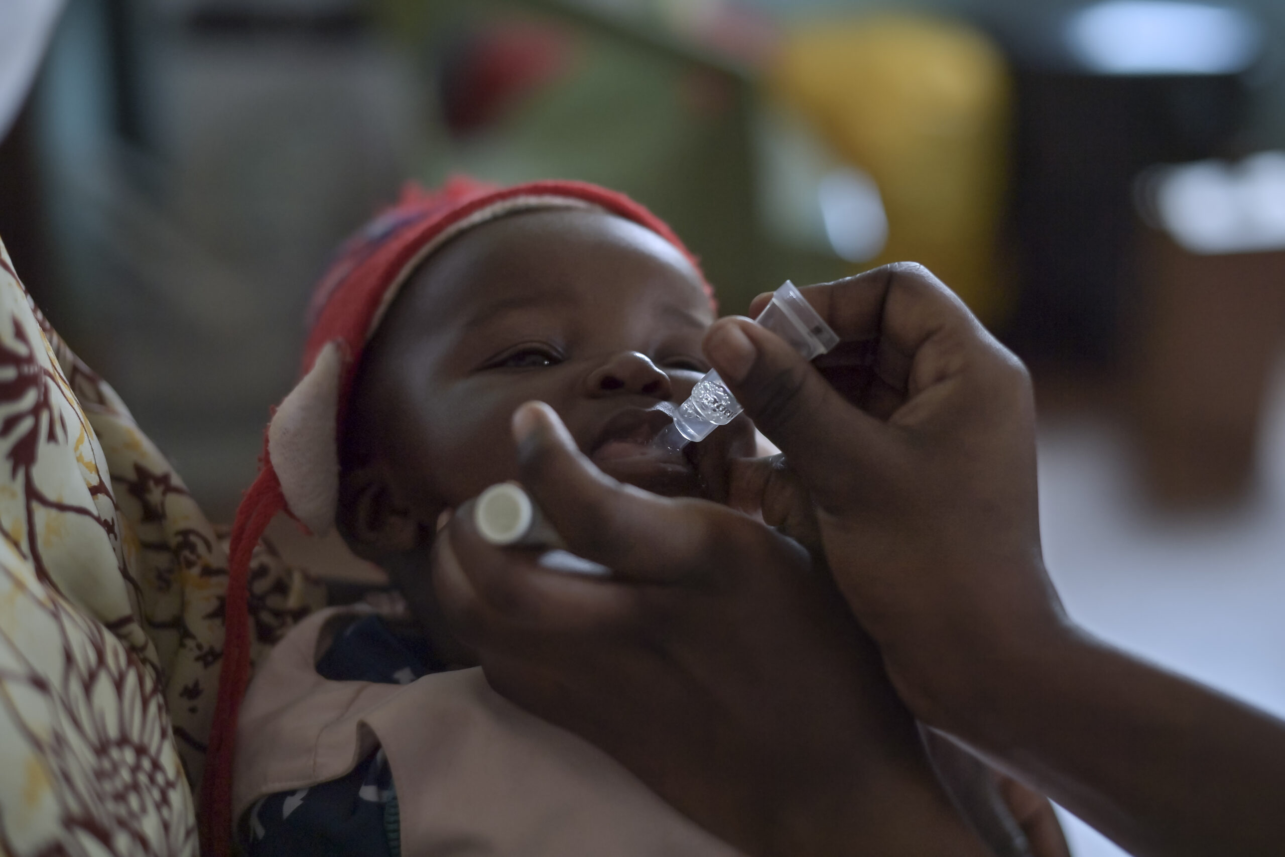 A close-up of a young child receiving an oral vaccine. The child is wearing a red and white hat, and two hands are gently administering the vaccine using a small dropper. The background is slightly blurred.