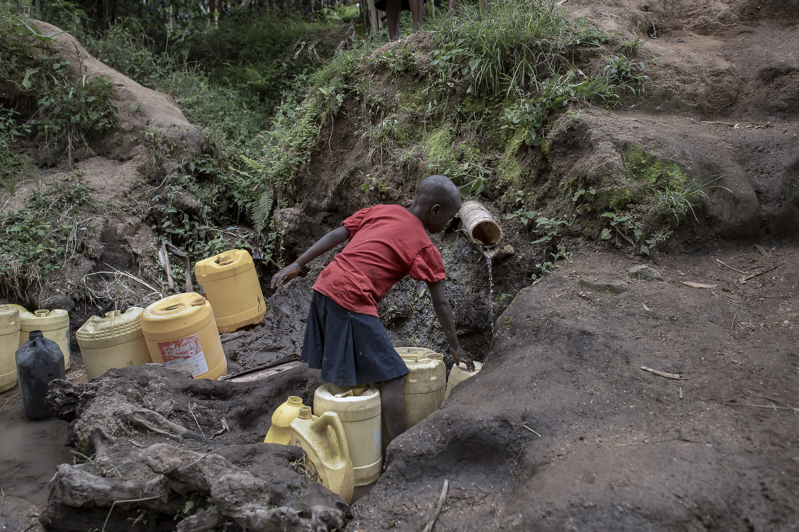 A child in a red shirt and dark shorts fills a yellow container with water from a small stream in a forested area. Several other yellow containers are placed on the ground around the child. The surroundings are muddy and covered in green foliage.