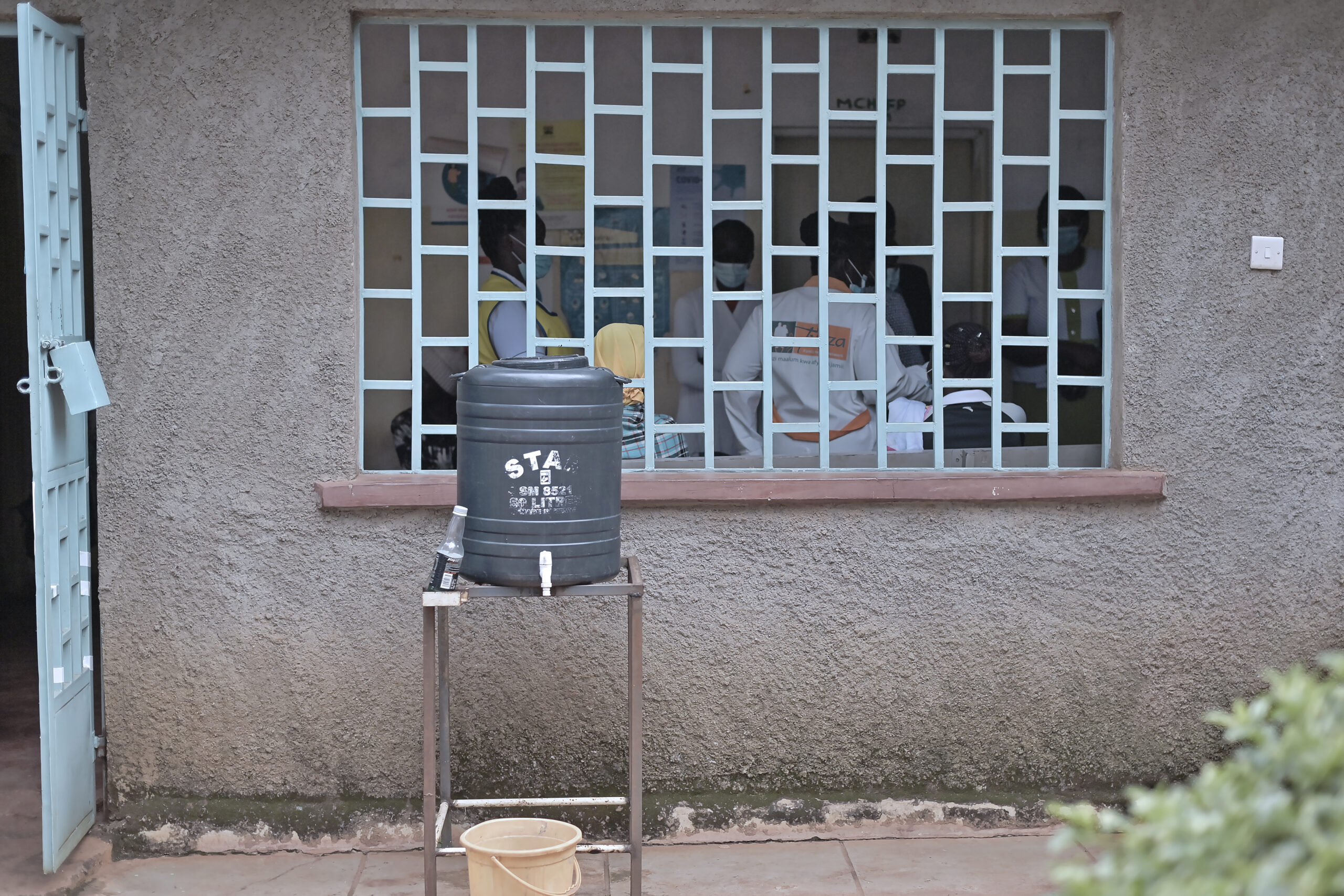 A handwashing station with a tank labeled "STA" stands outside a barred window of a building. Inside, several people in uniforms are visible, some wearing high-visibility vests. The scene suggests a healthcare or administrative setting.