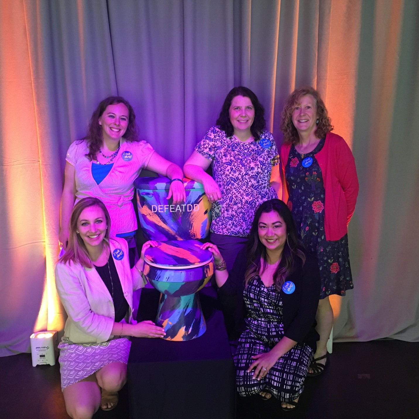 Five people are posing around a colorful toilet art piece. They are smiling and standing in front of flowy gray curtains under bright lighting. The word "DEFEATDD" is visible on the toilet. The group appears to be happy, vibrant, and supportive of a cause.