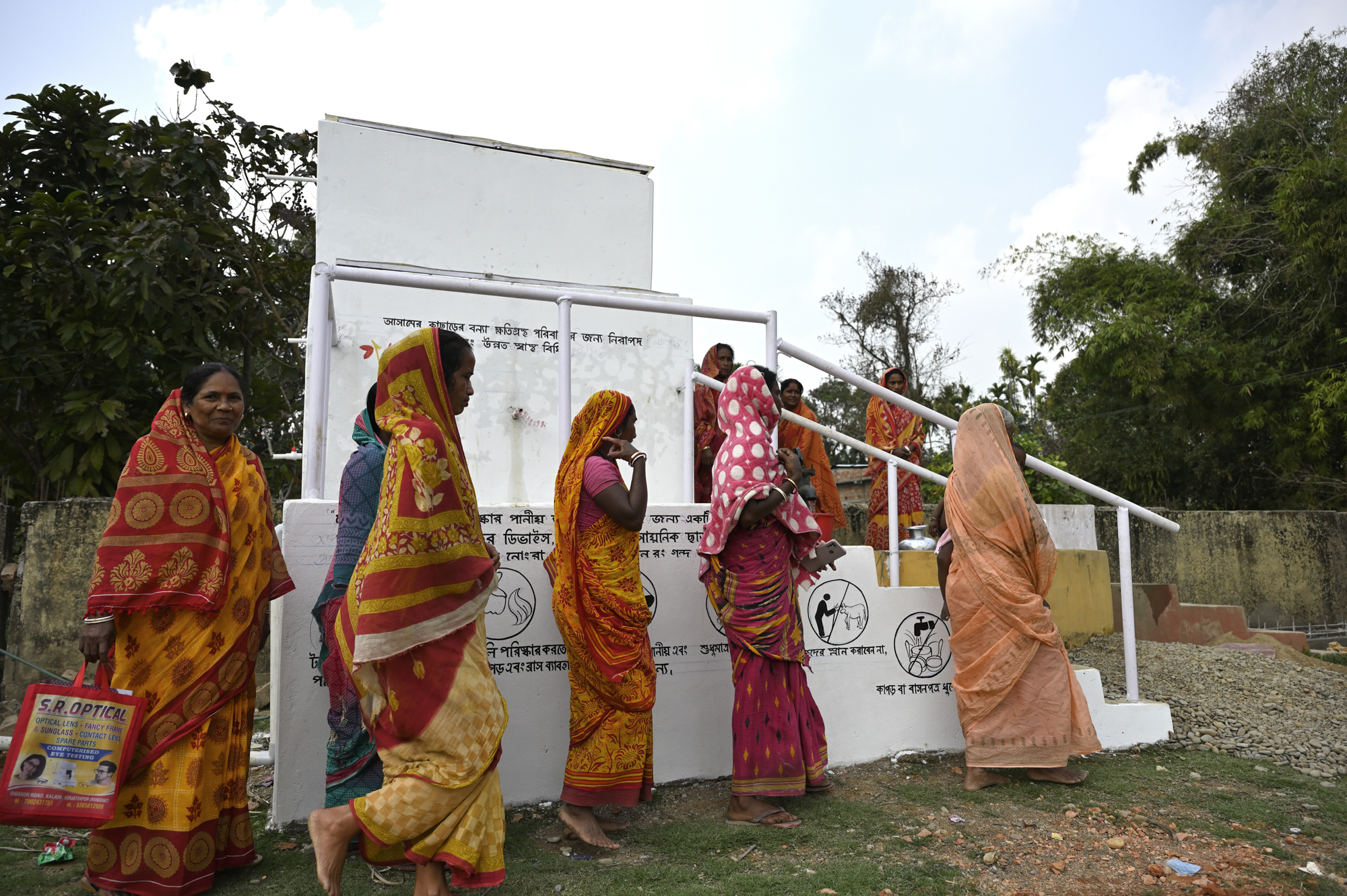 A group of women in colorful traditional clothing, some carrying bags, stand in line near a water facility. The background shows lush greenery and a white structure with signage and instructions written in a regional script. The sky is partly cloudy.