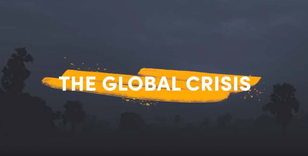 An image with a dark, cloudy background and silhouettes of trees. Overlaid in the foreground is a bold yellow paint stroke with the white text "THE GLOBAL CRISIS" written across it.