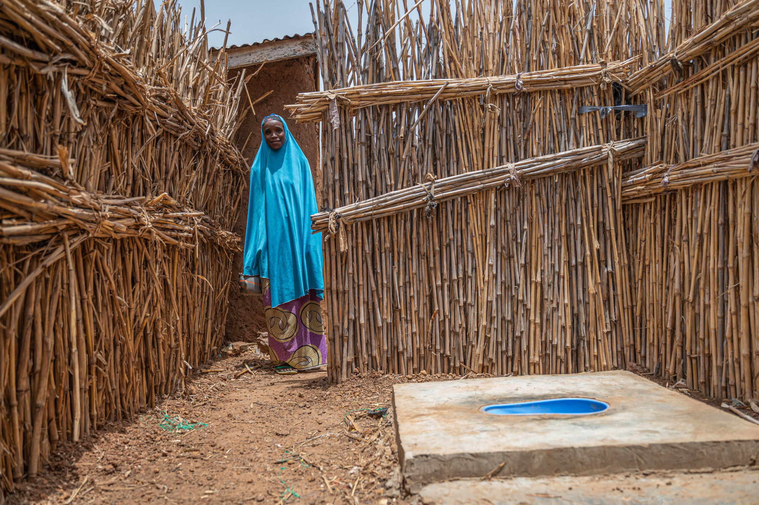 A person in a blue hijab stands outside behind walls made of vertical wooden sticks, next to a concrete slab with a blue-painted hole, which appears to be a latrine. The ground is bare and the walls create narrow pathways. The sky is grey.