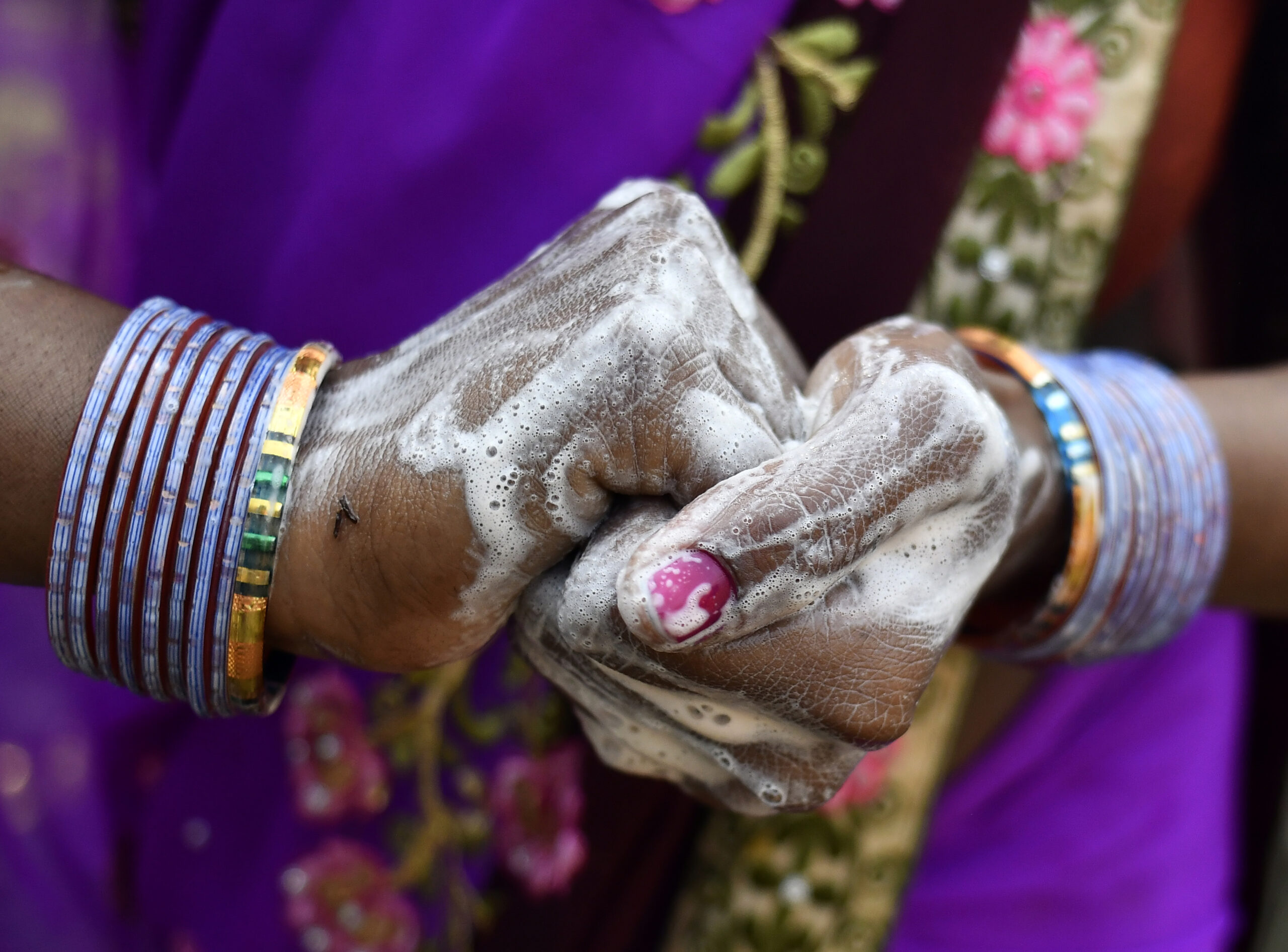 A close-up of a person's soapy hands clasped together. The person is wearing several colorful bangles and a purple sari with floral embroidery. The hands appear to be in the process of washing, with noticeable soap bubbles and lather.