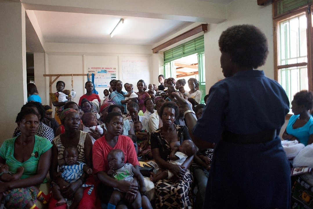 A healthcare worker addresses a room full of women and children in a community health center. Many women hold infants, and some children are seated beside their mothers. The room is crowded but orderly, with educational posters on the walls and windows providing natural light.