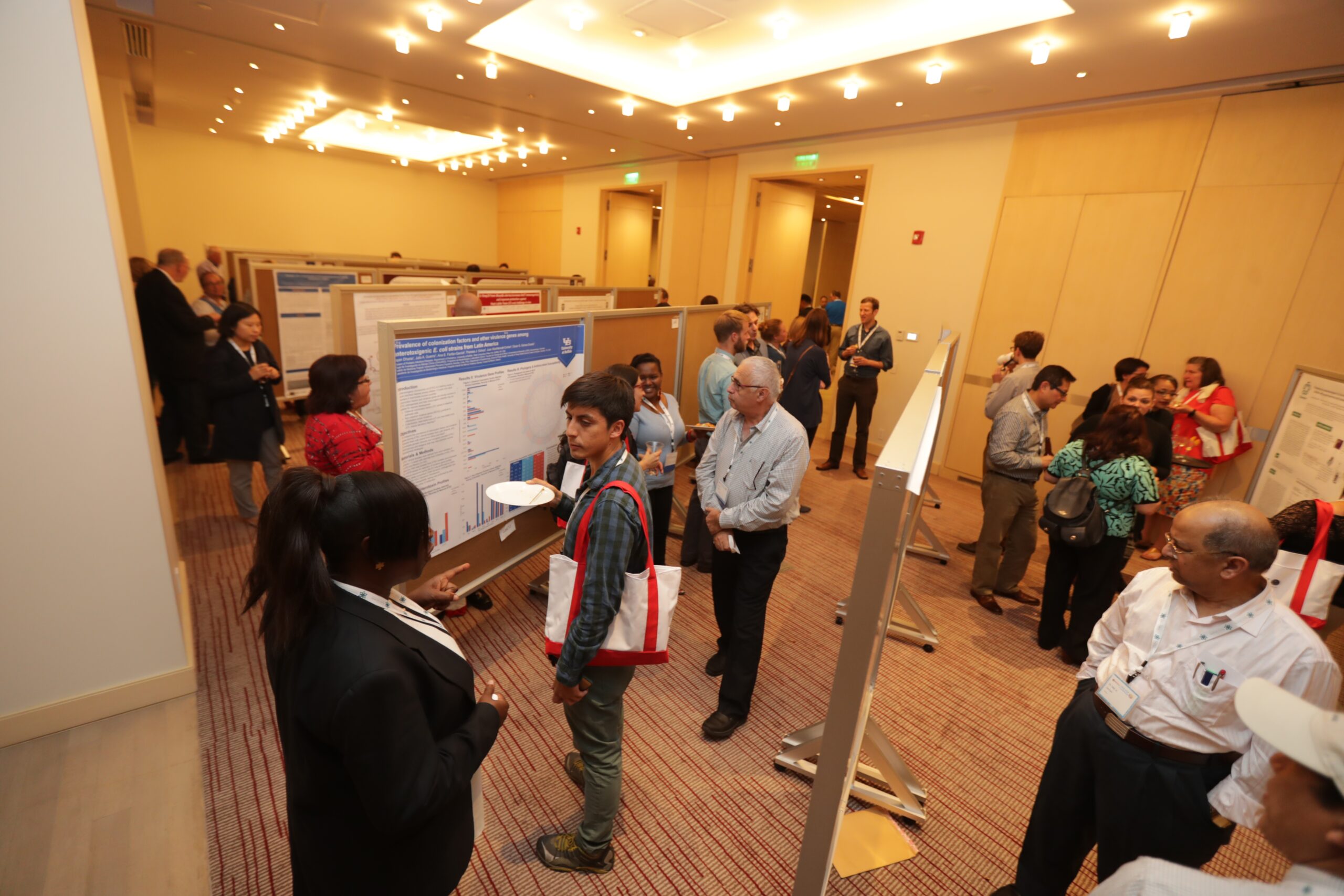 A conference room with people discussing research posters displayed on boards. Attendees are engaged in conversations, some holding tote bags and brochures. The room is well-lit with a carpeted floor and light-colored walls.