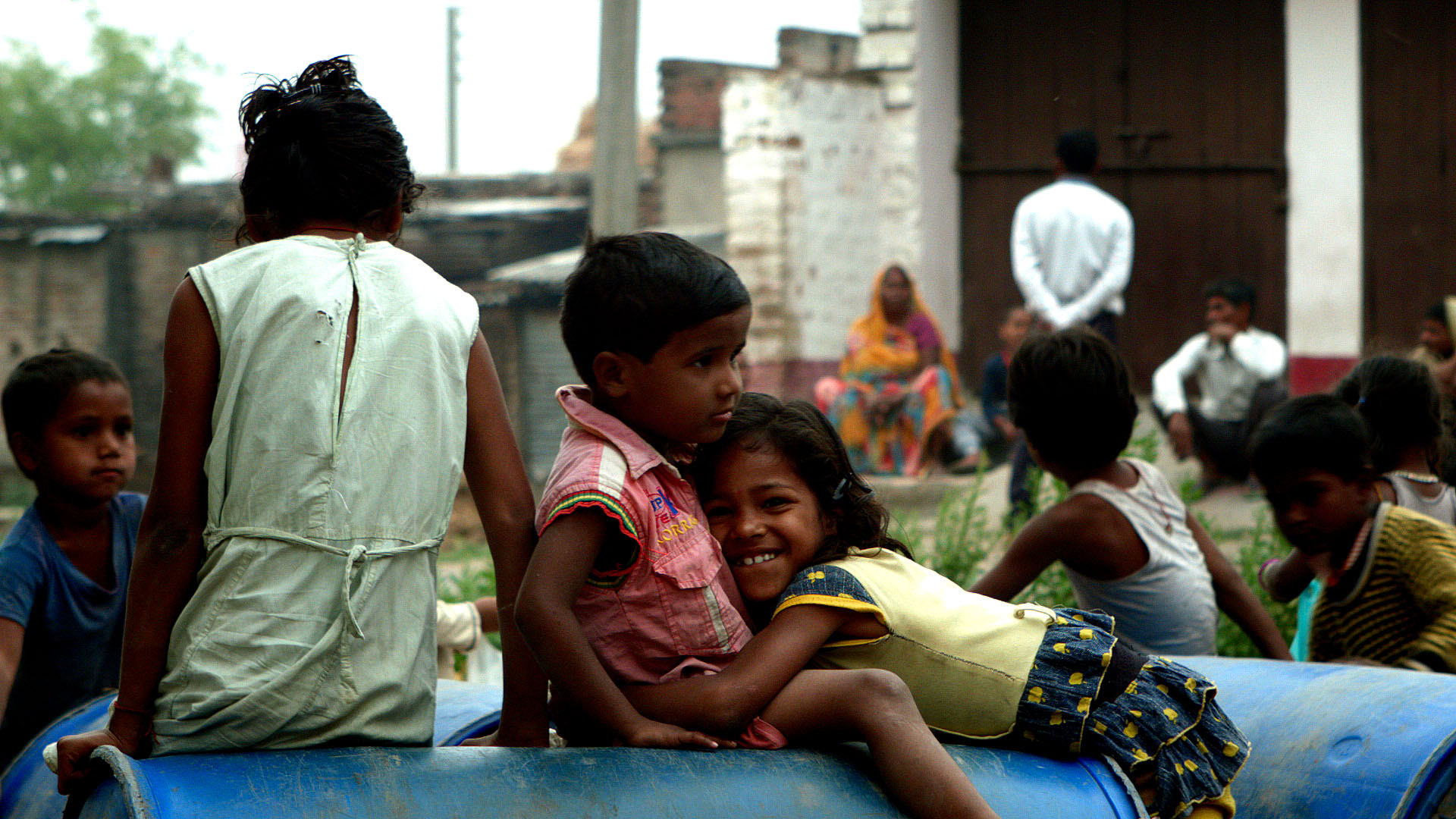 A group of children playing on large blue barrels in a rural setting. One child is smiling directly at the camera, and others are engaged in conversation. Adults and more children can be seen in the background near a brick building.