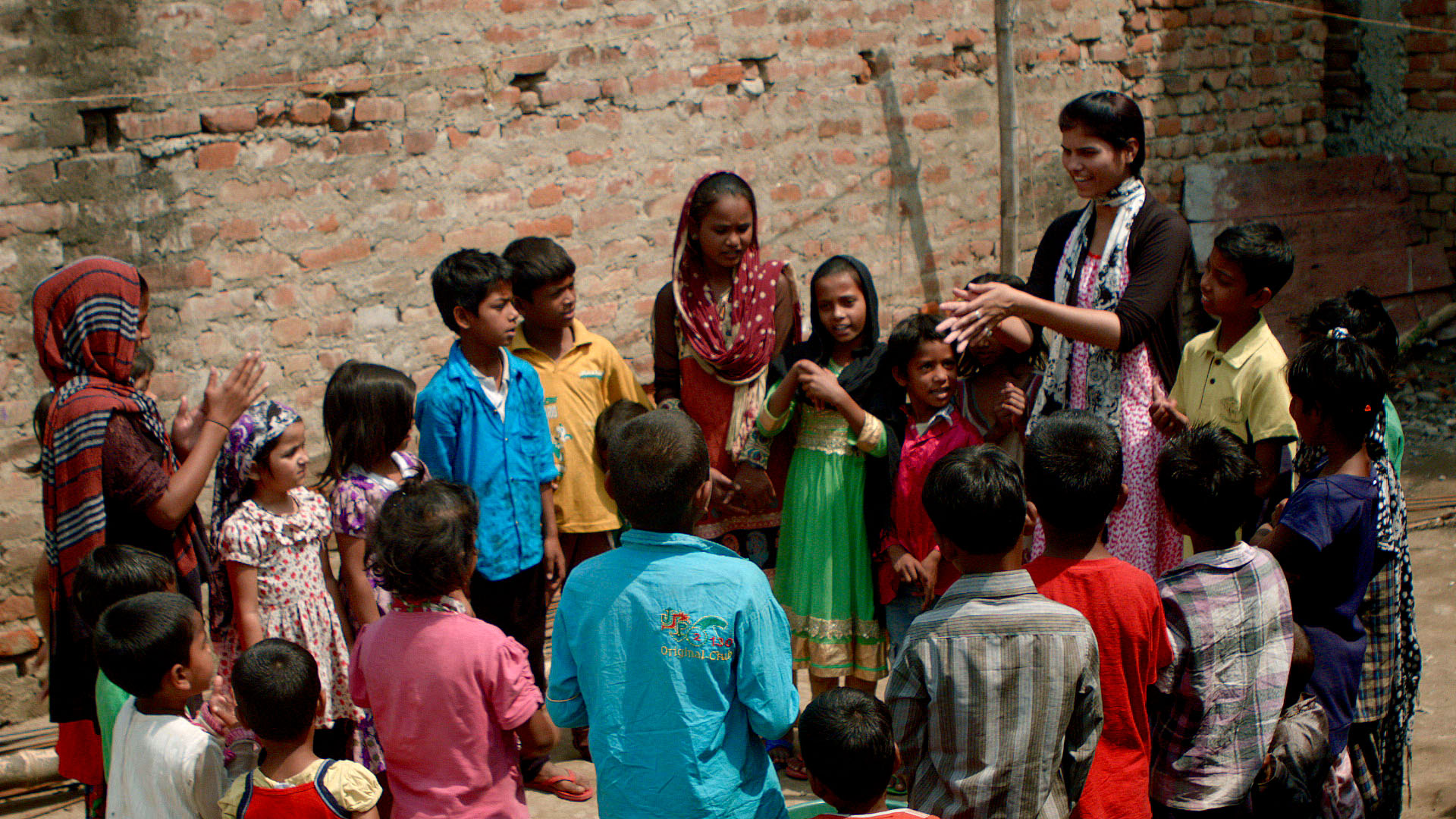 A group of children and two adults, all wearing colorful clothing, stand together in front of a brick wall. One adult appears to be speaking or instructing the children, who are listening attentively. The setting is outdoors, and the ground is uneven.